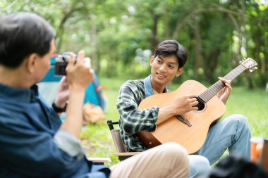 Bir babanın oğlunun gitar çalarken fotoğrafını çektiği, aile bağlarını ve açık hava eğlencesini vurguladığı hoş bir kamp sahnesi..