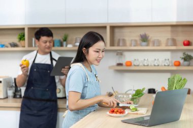 Arkadaşı elinde tablet tutarken bir kadın yemek hazırlıyor. Parlak bir mutfakta modern yemek tekniklerini sergiliyor..