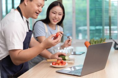 Bir çift, modern bir mutfakta taze domates ve salatalık hazırlarken dizüstü bilgisayarında yemek tarifleri araştırıyor..