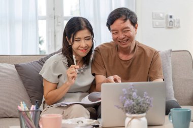 Yaşlı bir çift, aile gelirlerini ve giderlerini hesaplamak, mali planlama ve takım çalışmalarını desteklemek için bir dizüstü bilgisayar ve belgeler kullanarak kanepeye oturuyor..