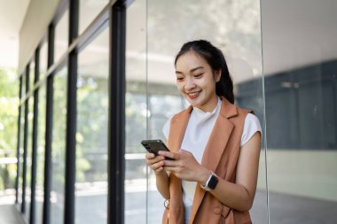 Genç bir kadın, parlak modern ofis ortamında akıllı telefonuyla iletişim kurarken gülümsüyor..