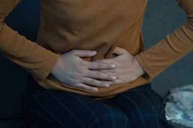 Oturan ve karnını tutan bir kişi, karın rahatsızlığını ve sağlık bilinci ihtiyacını gösterir..