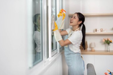 Önlüklü ve eldivenli neşeli bir kadın pencereleri temizliyor, ev temizliğine ve organizasyonuna kendini adamış..