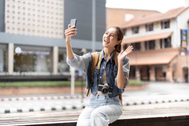 Genç bir kadın akıllı telefonuyla selfie çekerken gülümsüyor ve el sallıyor. Canlı bir şehirde neşeli anları yakalıyor..
