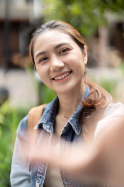 Genç bir kadın yemyeşil bir ortamda selfie çekerken gülümseyip el sallıyor, neşe ve bağlantı gösteriyor..