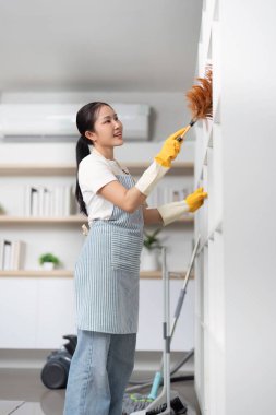 Genç bir kadın mutlu bir şekilde rafların tozunu alıyor. Temiz ve düzenli bir yuva kurma kararlılığını yansıtıyor..