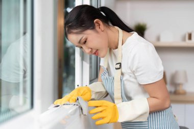 Sarı eldivenli bir kadın pencere çerçevesini inceliyor. Ev temizliğine olan bağlılığını gösteriyor..