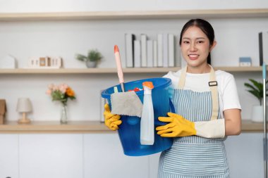 Neşeli bir kadın temizlik malzemeleriyle dolu mavi bir kovayla ayakta duruyor ve ev temizliği görevlerine hazır olduğunu gösteriyor..