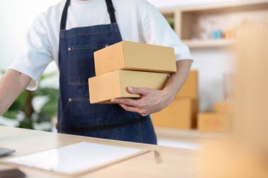 Önlüklü genç bir adam paketleri organize ediyor ve bilgisayarında çalışıyor, elektronik ticaretin ve uzaktan kumandanın dolandırıcılığını gösteriyor..