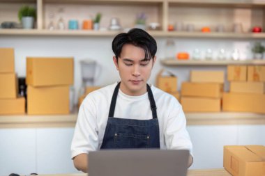 Önlüklü genç bir adam iyi organize edilmiş bir çalışma alanında online siparişleri yönetmek için bir dizüstü bilgisayar kullanıyor..