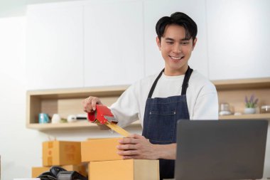 Neşeli bir işçi, modern e-ticaret sürecinin altını çizerek dizüstü bilgisayardaki siparişleri yönetirken kutuları bant ile mühürlüyor..