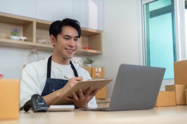 Önlüklü genç bir adam dizüstü bilgisayar üzerinde çalışırken paketler düzenler, uzaktan çalışma ve verimli lojistik yönetimini gösterir.