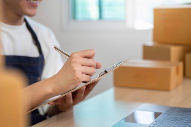 Odaklanmış bir e-ticaret çalışanı, bir dizüstü bilgisayarın siparişlerini yönetirken bir panoya notlar alır ve modern bir çalışma alanında verimliliği gösterir..