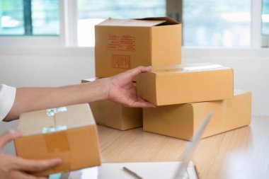 Genç bir adam karton kutularla dolu parlak bir ev ofisinde teslimat düzenlerken dikkatle bir paketi kaldırıyor..