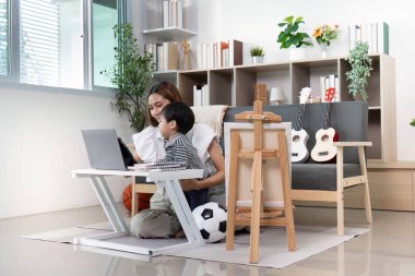 Bir anne ve oğlu, spor ve müzikal öğelerle dolu sıcak bir oturma odasında dijital öğrenmeyi teşvik eden bir dizüstü bilgisayarla uğraşıyorlar..