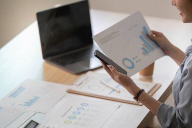 Profesyonel bir kadın akıllı telefonunu kontrol ederken kağıt üzerinde veri tablolarını analiz eder, modern iş uygulamalarını ve veri odaklı kararları yansıtır..