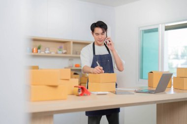 Genç bir adam, paketleri organize ederken ve not alırken, e-ticaretin hızını vurgularken, bir telefon görüşmesinde birden fazla iş yapar..