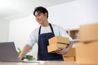 Neşeli genç bir adam, e-ticaretin ve uzaktan çalışmanın dinamik doğasını temsil eden paketler ve bir dizüstü bilgisayarı dengeler..