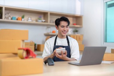 Önlüklü genç bir adam, ambalaj malzemeleriyle dolu modern bir çalışma alanında dizüstü bilgisayar kullanırken bir ürün tutuyor..