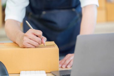 Önlüklü bir işçi, e-ticaret operasyonları ve lojistik faaliyetlerini gösteren yoğun bir nakliye bölgesinde bir paket üzerine yazıyor..