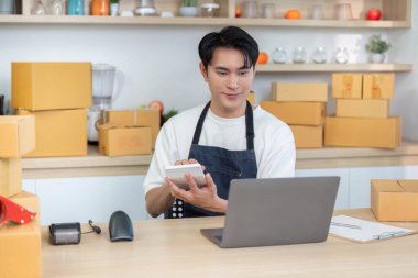 Önlüklü genç bir adam, iyi organize edilmiş bir ofis ortamında e-ticaret siparişlerini yönetirken tablet kullanıyor..