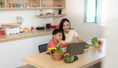 Neşeli bir anne ve oğlu, sıcak mutfaklarında dizüstü bilgisayar kullanırken eğlenceli bir yemek pişirme deneyimini paylaşıyorlar. Aile bağlarını destekliyorlar..
