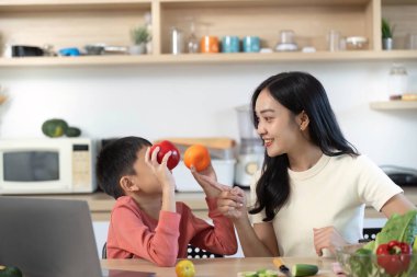 Bir anne ve oğlu, elmalarla portakalları tutarak, mutfakta sağlıklı beslenme ve aile bağlarını vurgulayarak, neşeli bir anı paylaşıyorlar..