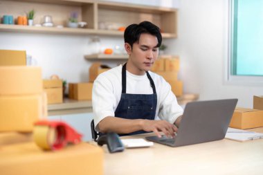 Önlüklü genç bir adam dizüstü bilgisayarda çalışıyor, e-ticaret siparişlerini kutularla dolu düzenli bir çalışma alanında işliyor..