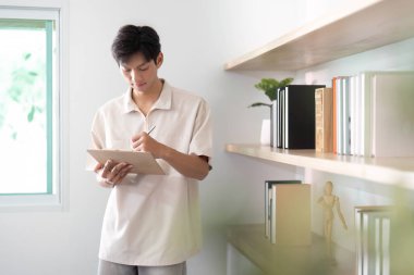 Genç bir adam modern bir ev ofisinde duruyor, bir panoya notlar yazıyor, uzaktan çalışma ve üretkenlik eğilimini yansıtıyor..