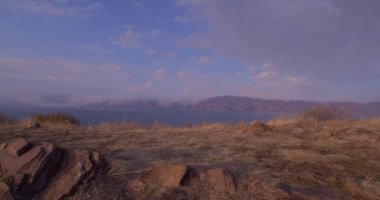 Ermenistan dağları arasında Sevan Gölü Panoraması. Bir köy yolu. Turistler geliyor. Keskin taşlar ve kayalar görülebilir. Bulutlar dağlara düşüyor. Turuncu çimenler sallanıyor