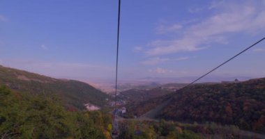 Ermenistan dağları arasında bir teleferik gezisi. Çöl kulübesi buradan geçiyor. Yeşil ve kırmızı ağaçlar yüzüyor. Sonbahar. Uzak dağlara ve şehre. Mavi gökyüzü. Bulutlar süzülüyor