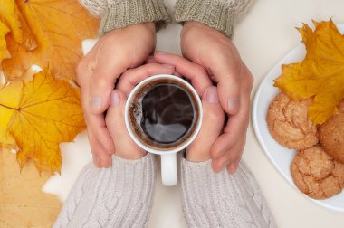 Sonbahar sarısı akçaağaç yapraklarının üst görüntüsü, erkeğin elleri kadının ellerinin üzerinde sıcak kahve fincanı tutmalı, fildişi arka planda kurabiyeler olan beyaz tabak. Sonbaharda rahat ev kavramı