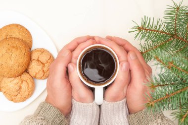 En tepedeki yeşil dallar, zencefilli kurabiyeli beyaz tabak, bir erkeğin elleri kadının ellerinde fildişi rengi arka planda bir fincan sıcak kahveyi tutmalı. Kışın ve Noel tatillerinde rahat ev kavramı