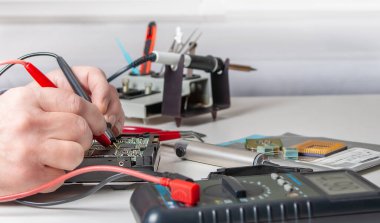 Atölyede bir teknisyenin ellerine yakın çekim. Tamirci, masadaki elektronik cihazın parametrelerini ölçmek için çoklu metre kullanıyor..