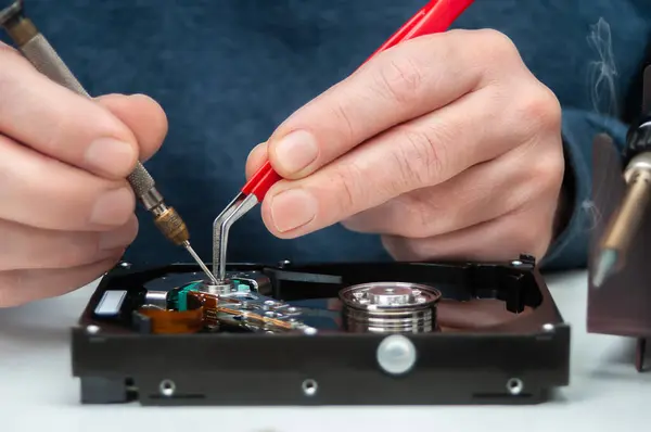 Close-up of a technician's hands in a workshop. The technician is using tweezers and a screwdriver to repair and maintain a hard drive.