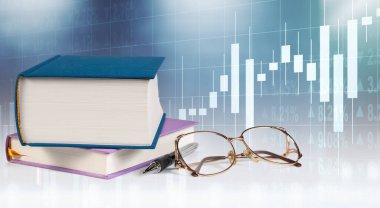 Ofis malzemelerinin yanına, kalemler ve bardaklar da dahil olmak üzere bir kitap koleksiyonu yığılır. Hepsi de aydınlatılmış bir borsa grafiğinin arka planına karşıdır..