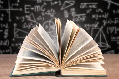 Açık bir ders kitabı, çeşitli matematik denklemleri ve diyagramlarıyla kaplı bir karatahtanın yer aldığı bulanık bir arkaplan ile ön plandaki ahşap bir masada yatar..