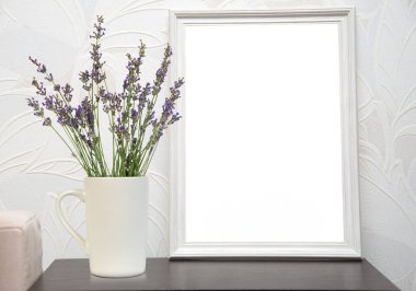 Lavanta çiçekli beyaz bir fincan beyaz bir resim çerçevesinin yanında bir masada oturur. Arka plan kabartmalı desenli beyaz bir duvar..