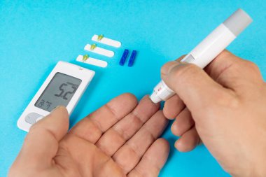 Bir el, glukometre ve mavi arka planda test şeritleriyle glikoz testi için kan almaya hazırlanıyor..