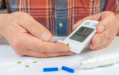 Kan şekeri ölçümlerini gösteren bir glukometre tutarken test bantları yakınlarda görünüyor. Bu da ev sağlığı yönetimini gösteriyor..