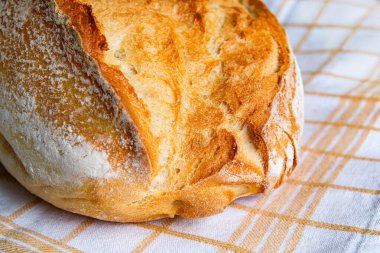 Yuvarlak bir somun ekmek, kabuklu ve altın sarısı, sıcak bir mutfakta keten bir kumaşın üzerinde oturur, sıcak bir atmosferi davet eder..