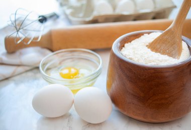 Bir mutfak tezgahında yumurta ve un koleksiyonu, çırpı ve oklavayla birlikte, pişirme faaliyetlerini bekliyor..