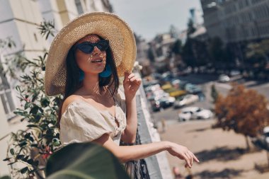 Rahat genç bir kadın balkonda dururken zarif şapkasını düzeltiyor.