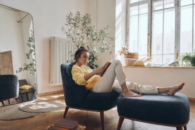 Evde rahat bir sandalyede rahatlarken kitap okuyan çekici genç bir kadın.