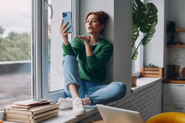 Güzel bir kadın akıllı bir telefon tutuyor ve pencerede otururken bir öpücük yolluyor.