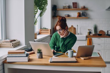 Evde mutfak tezgahında otururken dizüstü bilgisayarda çalışan güzel genç bir kadın.
