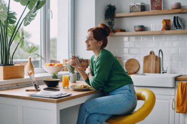 Evdeki mutfak tezgahında oturmuş öğle yemeği yiyen hayalperest genç kadın.