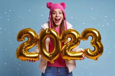 Kışlık ceketli güzel bir kadın altın renkli numaralar taşıyor ve mavi arka planda gülümsüyor.