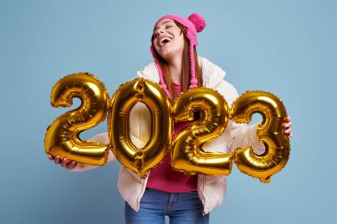 Mavi arka planda altın sarısı numaralar taşıyan kışlık ceketli mutlu genç kadın.