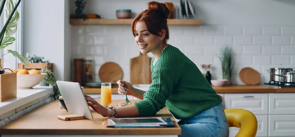 美しい若い女性が昼食を取り キッチンカウンターに座っている間にデジタルタブレットを使用して — ストック写真